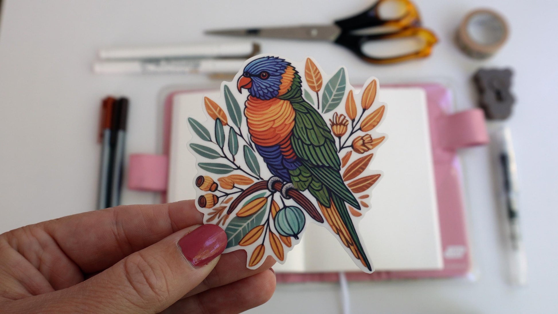 A hand holds a sticker of a colorful rainbow lorikeet perched on a branch with leaves and buds. In the background, an open notebook with a pink cover sits on a white surface along with scattered pens, scissors, and a roll of tape.
