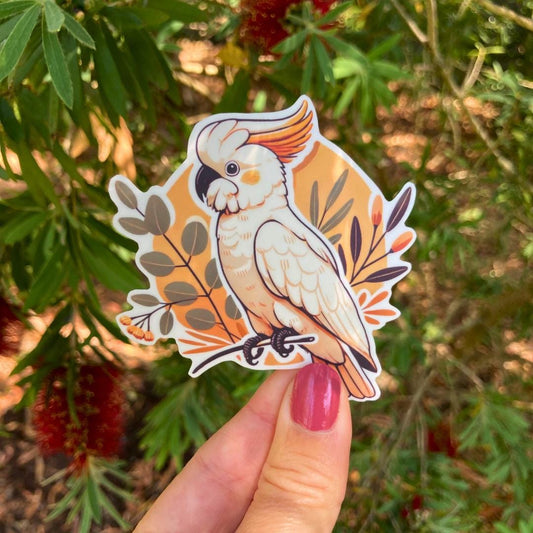 A hand holds a sticker of a white cockatoo perched on a branch. The bird has an orange and yellow crest, surrounded by a background of orange and green leaves. A leafy tree is blurred in the background. The person’s nails are painted pink.