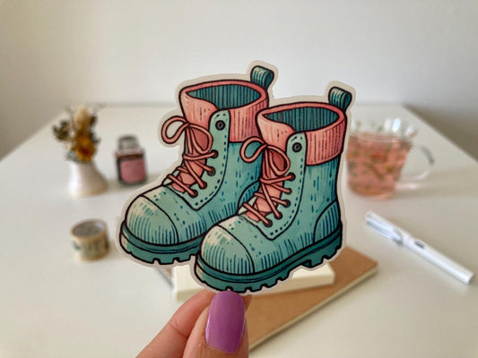 A hand with purple nails holds a garden boots sticker featuring teal boots with pink laces and cuffs. The background shows a desk with a notebook, dried flowers, a tea cup, and stationery.