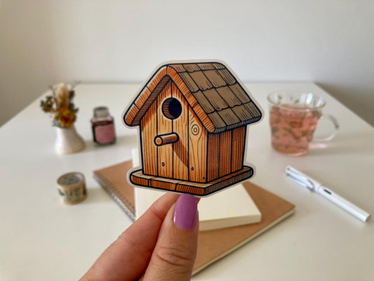 A hand with purple nails holds a birdhouse sticker depicting a wooden house with a round entrance and shingled roof. The background shows a desk with a notebook, dried flowers, a tea cup, and stationery.
