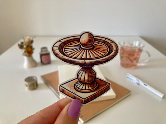 A hand with purple nails holds a birdbath sticker featuring a decorative pedestal and round basin. The background shows a desk with a notebook, dried flowers, a tea cup, and stationery.