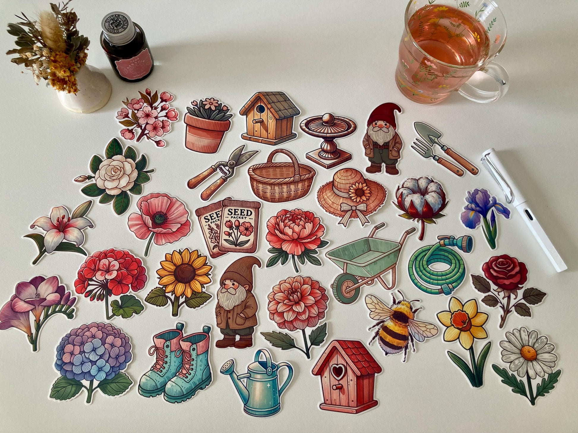 A collection of garden-themed stickers, including flowers, gardening tools, seed packets, gnomes, a birdhouse, boots, a watering can, a wheelbarrow, and a bee. Nearby are dried flowers, a tea cup, a white pen, and an ink bottle on a white table.
