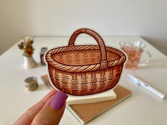 A hand with purple nails holds a wicker basket sticker featuring a curved handle and woven design. The background shows a desk with a notebook, dried flowers, a tea cup, and stationery.