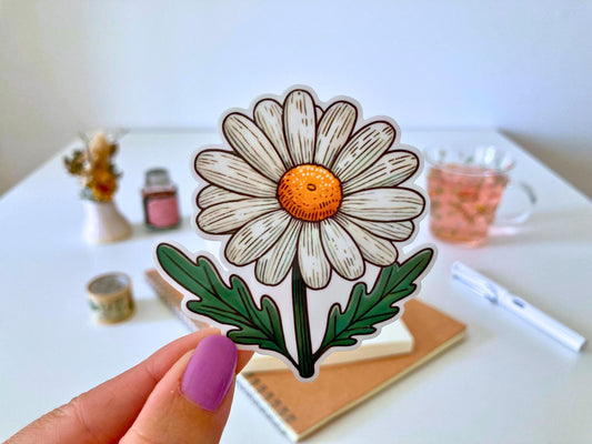 A hand with purple nails holds a daisy sticker featuring white petals, a yellow center, and green leaves. The background shows a desk with a notebook, dried flowers, a tea cup, and stationery.