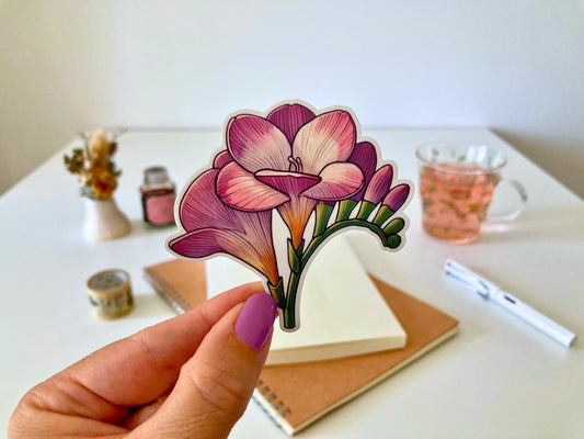 A hand with purple nails holds a freesia sticker featuring pink and purple trumpet-shaped flowers with green stems and buds. The background shows a desk with a notebook, dried flowers, a tea cup, and stationery.