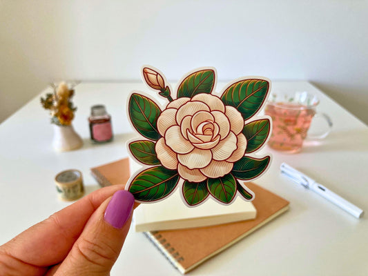 A hand with purple nails holds a camellia sticker featuring layered cream petals, green leaves, and a closed bud. The background shows a desk with a notebook, dried flowers, a tea cup, and stationery.