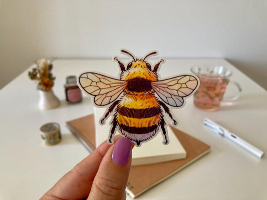 A hand with purple nails holds a detailed bee sticker with yellow and black stripes and translucent wings. The background shows a desk with a notebook, dried flowers, a tea cup, and stationery.