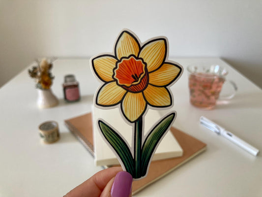 A hand with purple nails holds a daffodil sticker with yellow petals, an orange trumpet, and green leaves. The background shows a desk with a notebook, dried flowers, a tea cup, and stationery.