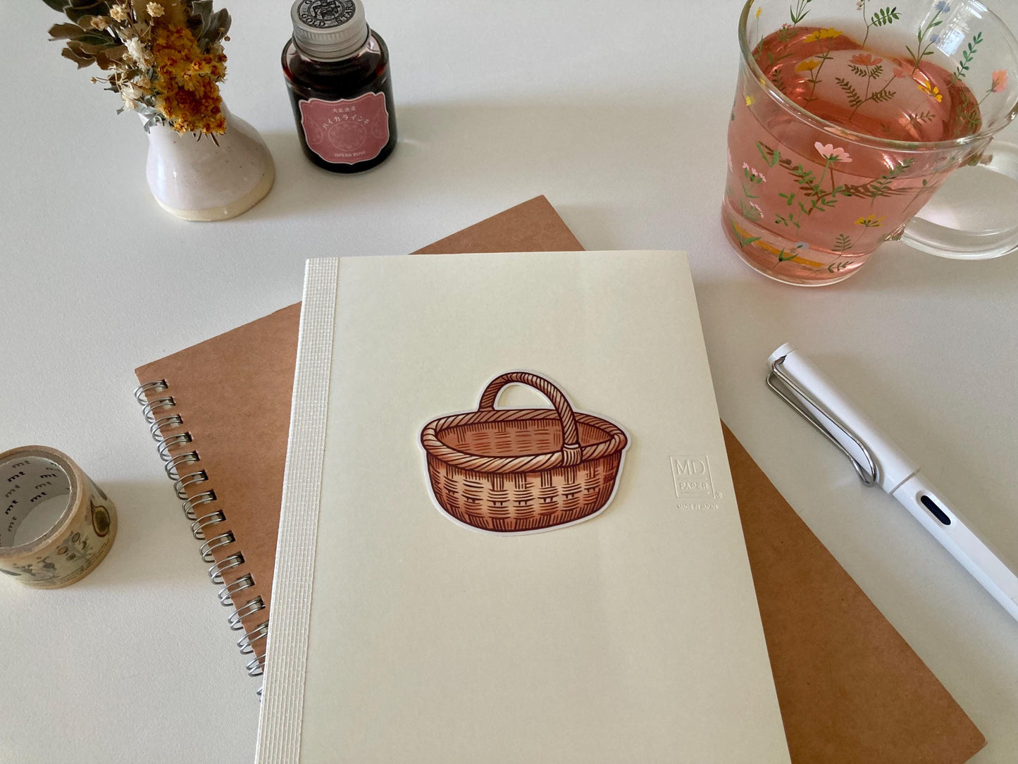 A wicker basket sticker with a curved handle is placed on a cream-colored Midori notebook atop a brown spiral notebook. Nearby are dried flowers, a tea cup, washi tape, an ink bottle, and a white pen.
