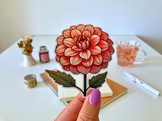 A hand with purple nails holds a dahlia sticker featuring layered red and cream petals with green leaves. The background shows a desk with a notebook, dried flowers, a tea cup, and stationery.