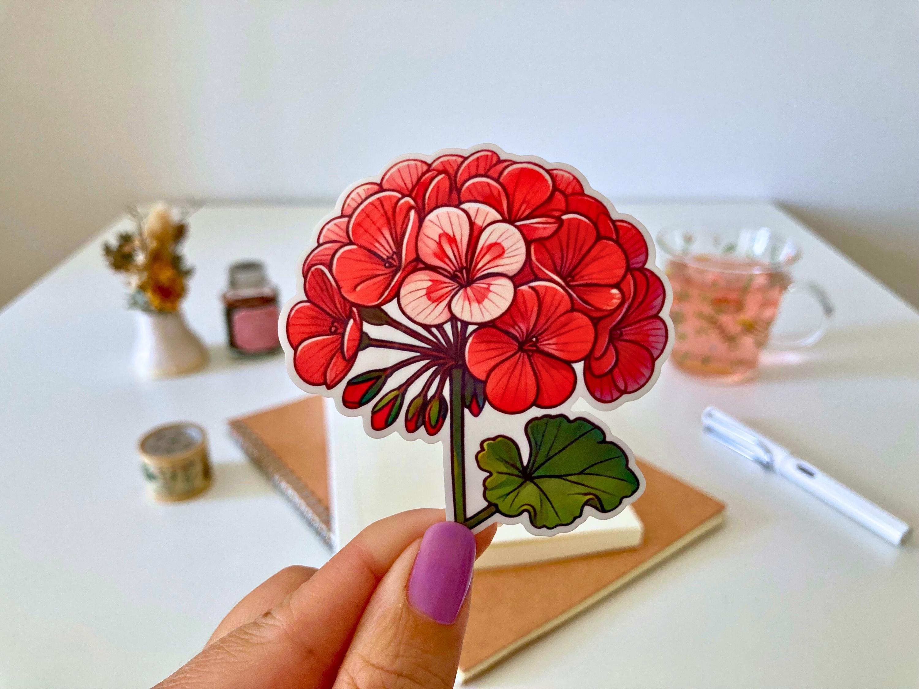 A hand with purple nails holds a geranium sticker featuring vibrant red and pink flowers with green, rounded leaves. The background shows a desk with a notebook, dried flowers, a tea cup, and stationery.