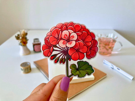 A hand with purple nails holds a geranium sticker featuring vibrant red and pink flowers with green, rounded leaves. The background shows a desk with a notebook, dried flowers, a tea cup, and stationery.