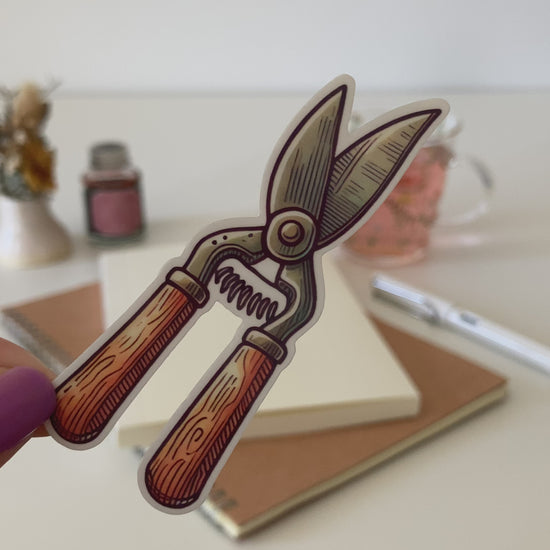 A hand with purple nail polish holds a cartoon sticker of vintage-style garden secateurs with wooden handles. The background features a desk with a notebook, pen, dried flowers, washi tape, a jar, and a glass cup of tea.
