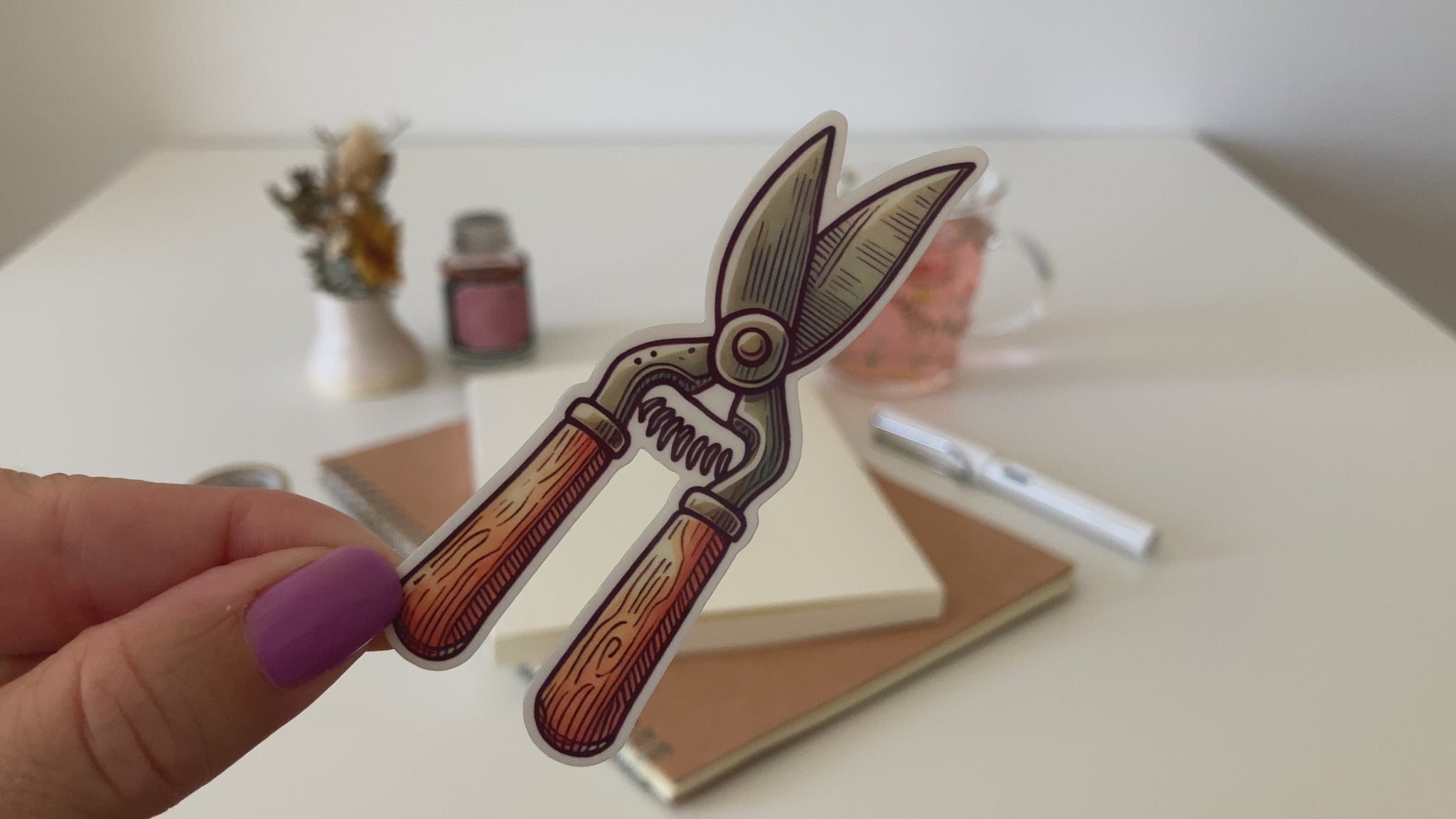 A hand with purple nail polish holds a cartoon sticker of vintage-style garden secateurs with wooden handles. The background features a desk with a notebook, pen, dried flowers, washi tape, a jar, and a glass cup of tea.