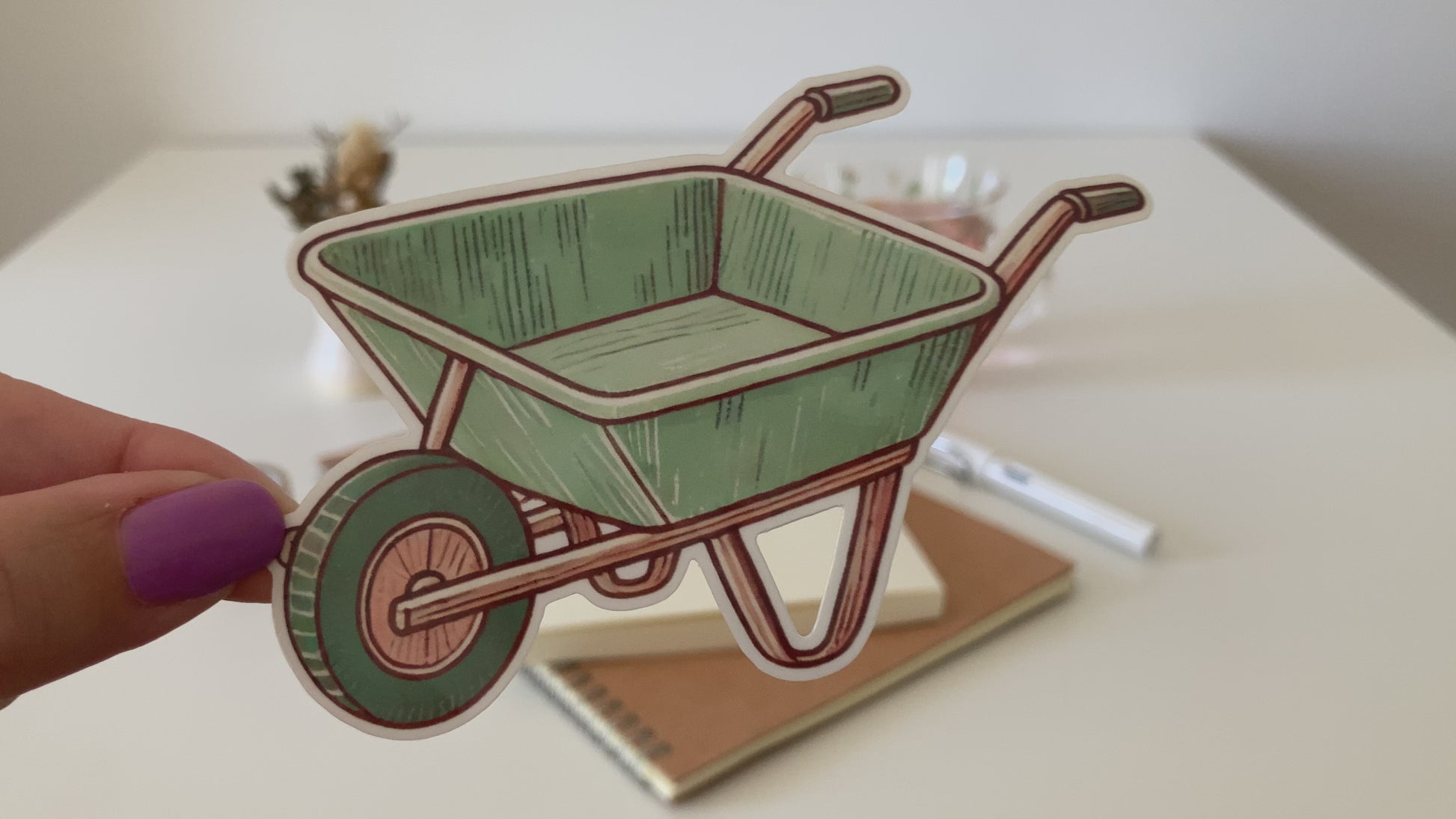 A hand with purple nail polish holds an anime-style sticker of a green wheelbarrow with brown outlines. The background features a white table with notebooks, a pen, a glass of pink tea, and decorative items.