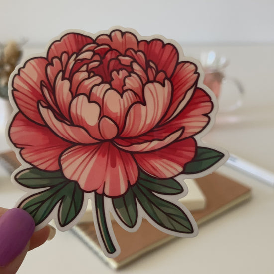 A hand with purple-painted nails holds a cartoon-style peony sticker, featuring layered red and pink petals with green leaves. The background shows a white table with dried flowers, a tea cup, a notebook, and a pen.