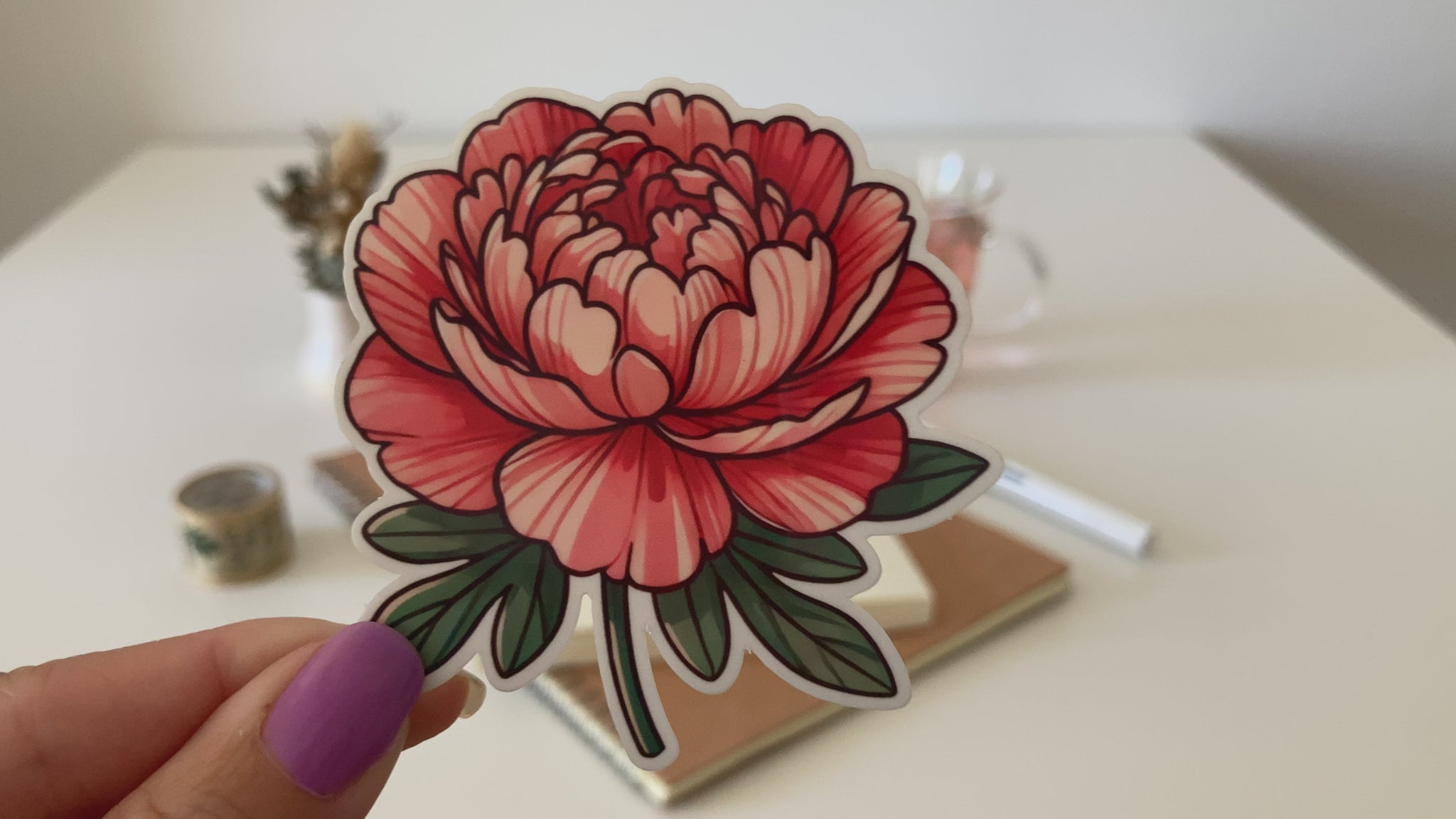 A hand with purple-painted nails holds a cartoon-style peony sticker, featuring layered red and pink petals with green leaves. The background shows a white table with dried flowers, a tea cup, a notebook, and a pen.